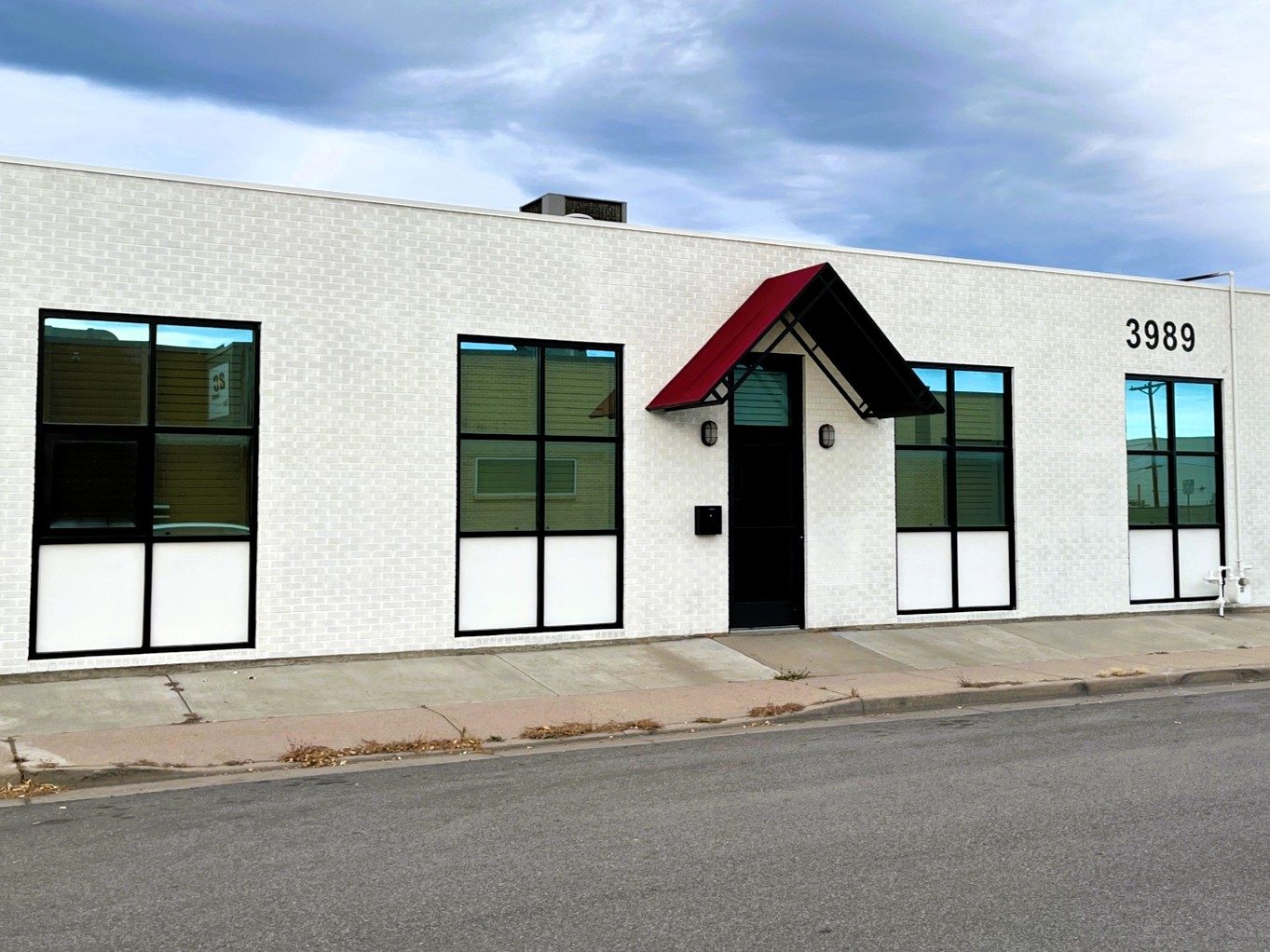 The white masonry front of 3989 S Lipan Street from across the street, with tall black-framed windows and a red awning over the entrance.